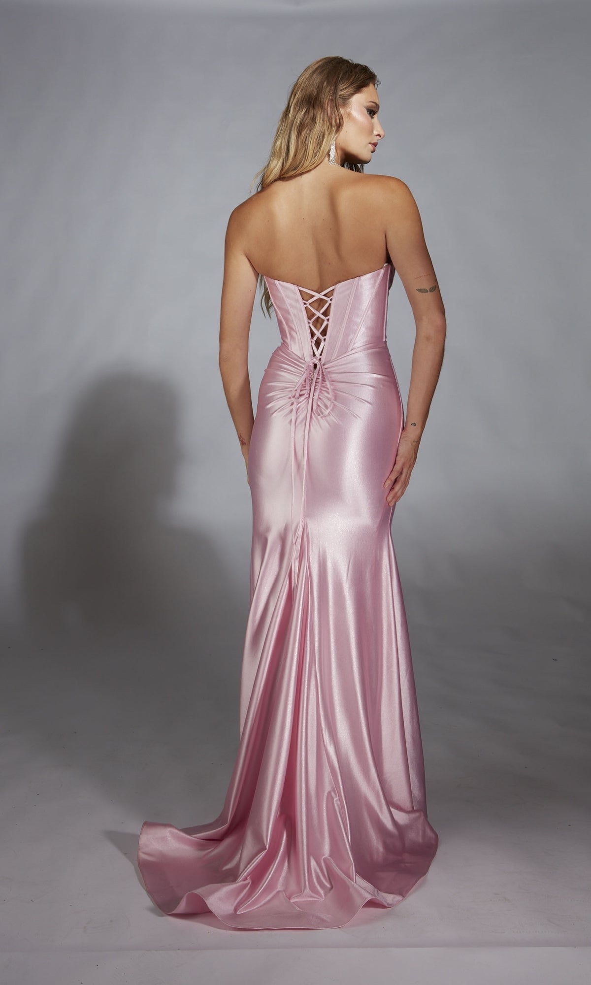 A woman in the Alyce 61733 Strapless Long Corset Prom Dress, a shiny pink gown with a lace-up back, stands facing away against a plain gray background.